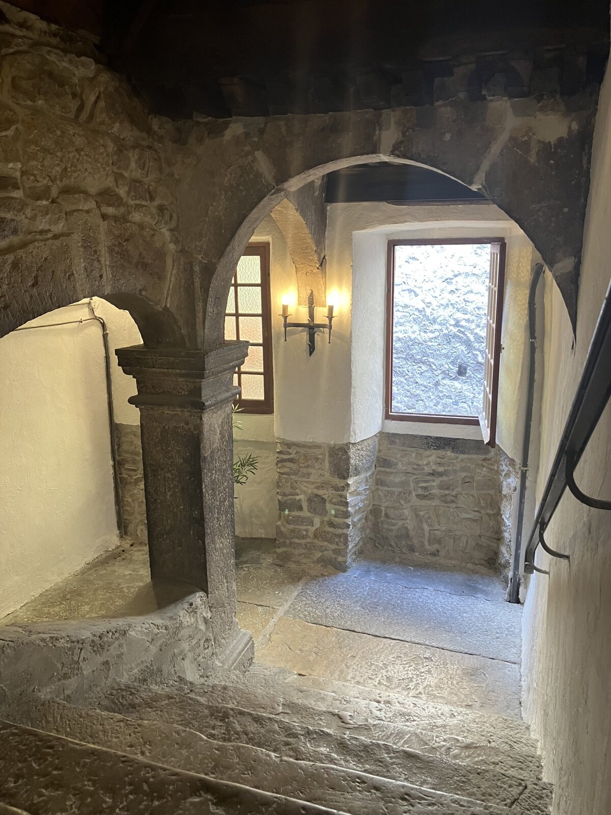 An interior stairway is highlighted, featuring a stone staircase with arched openings. Natural light floods in from a window, illuminating the textured stone walls and wooden beams above. A wrought iron sconce adds a touch of historical charm, complementing the rustic ambiance.