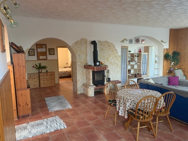A spacious living area features a rustic fireplace surrounded by stonework. A wooden dining table with chairs is positioned nearby, and a cozy sofa with decorative cushions invites relaxation. The room has a warm ambiance, complemented by large openings leading to other areas.
