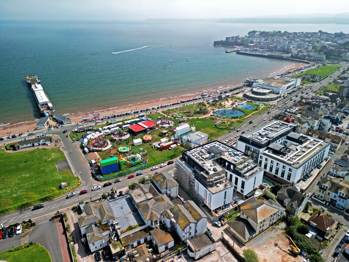 An aerial view captures the vibrant coastline of Paignton, showcasing the sandy beach and the lively promenade lined with attractions. Colorful structures, including amusement rides and food stalls, are visible on the green expanse, while the calm sea reflects the sky, providing a scenic backdrop.