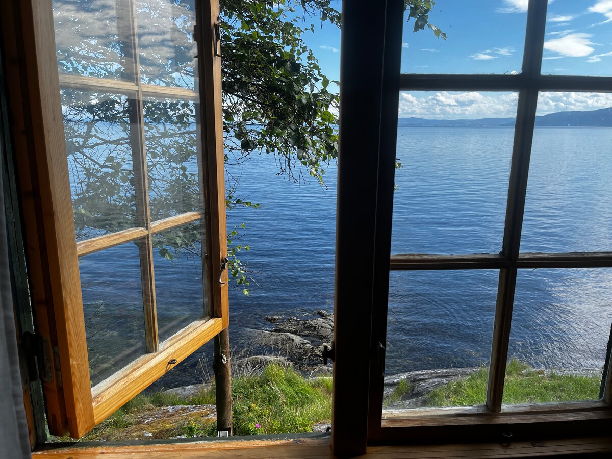 A wooden window is open, framing a view of the tranquil water below. The calm surface reflects the clear blue sky, while greenery is visible just outside the window. The scene captures the serene proximity to nature.