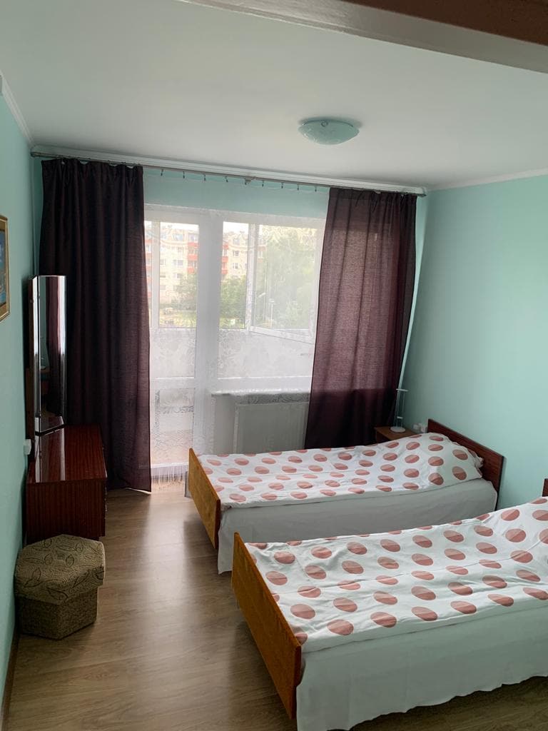 The bedroom features two twin beds with polka dot bedding. A large window with dark curtains allows natural light to fill the room, while a small ottoman sits beside the beds. A wooden dresser is positioned against one wall.