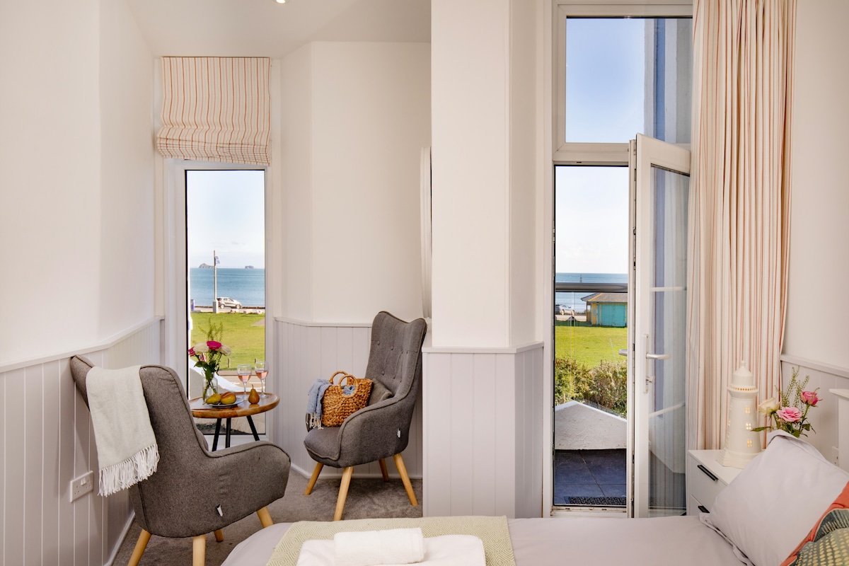 A light-filled studio apartment features two armchairs beside a small table, with views of the sea visible through large windows. Soft furnishings in neutral tones complement the calming atmosphere, while a glass door opens to an outdoor area, enhancing the seaside experience.