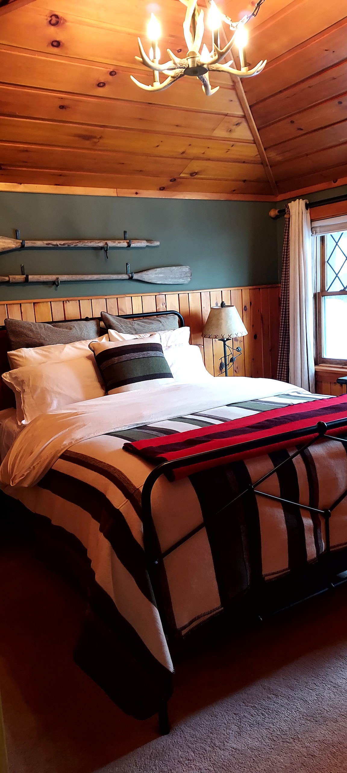 The bedroom features a cozy queen bed adorned with luxury bedding. Natural wood paneling and a vaulted ceiling enhance the warm atmosphere. A bedside lamp sits next to the bed, and a large window allows for natural light. Decorative oars are mounted on the wall.