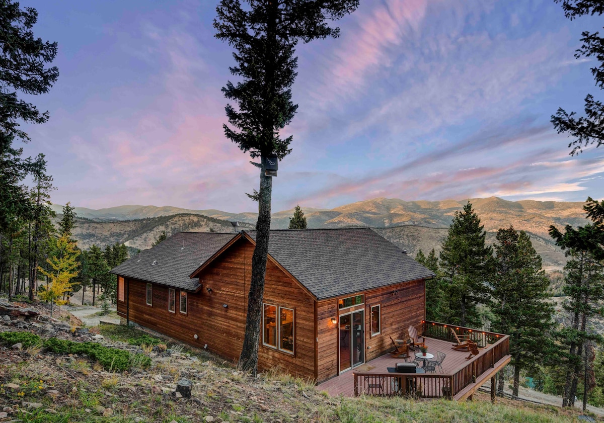 A custom-built wooden cabin is set against a backdrop of mountains and colorful sky at dusk. The spacious deck showcases outdoor seating, while tall trees surround the home, enhancing its natural charm in a serene mountain landscape.