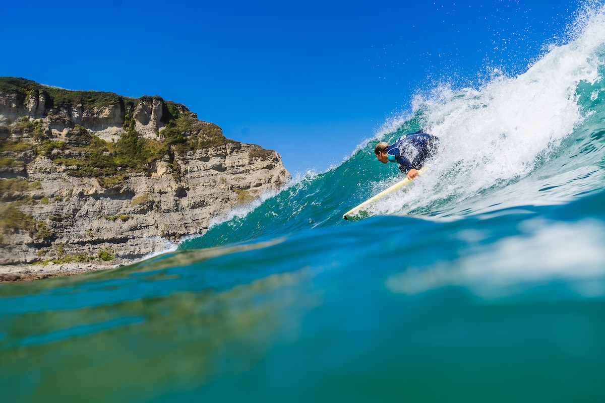 A surfer is captured riding a wave in clear turquoise water. The image features a stunning backdrop of rugged cliffs under a bright blue sky, highlighting the natural beauty of the surfing location.