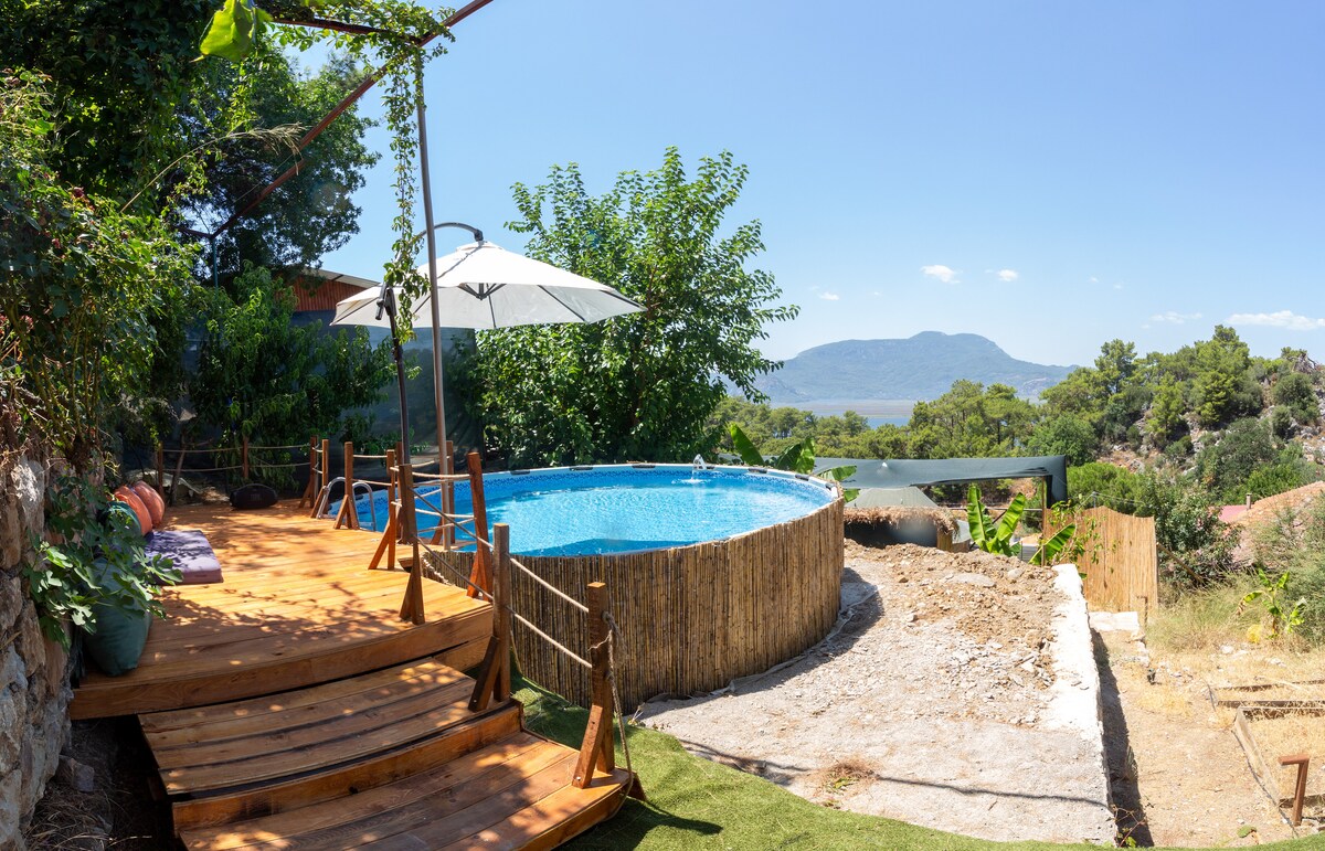 A circular above-ground pool is set within a natural landscape, surrounded by lush greenery and distant hills. Wooden decking offers a welcoming space for relaxation, complemented by a large umbrella providing shade. The tranquil setting invites outdoor enjoyment and peaceful moments.