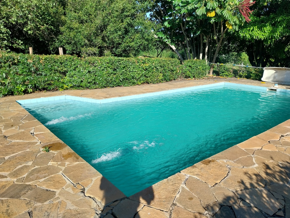 A refreshing swimming pool is set within a stone patio, surrounded by lush greenery. The water displays a vibrant turquoise hue, with gentle waves reflecting sunlight. Trees and bushes provide a natural border, creating a tranquil outdoor space for relaxation.