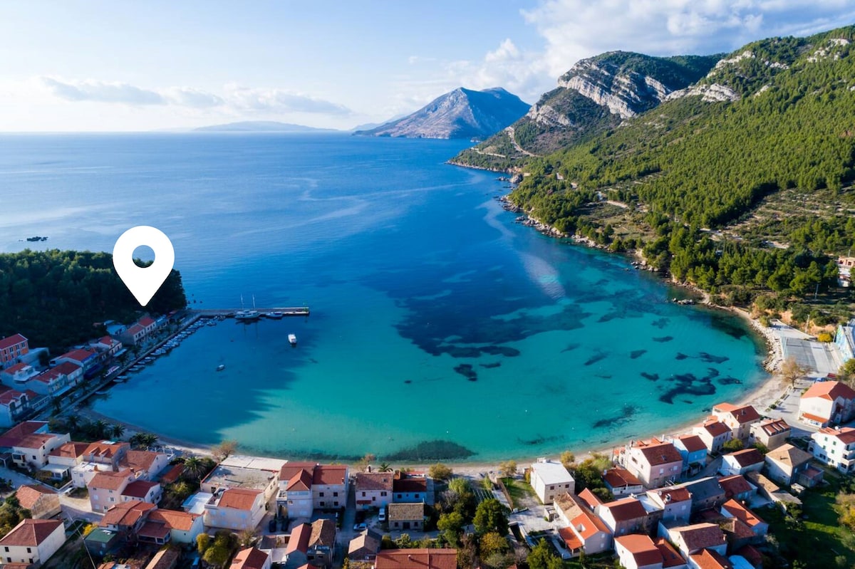 An aerial view showcases a serene bay surrounded by lush hills and clear blue waters. The coastline displays charming buildings with terracotta roofs. A small marina is visible, with boats gently bobbing in the water. Scenic hills rise in the background.