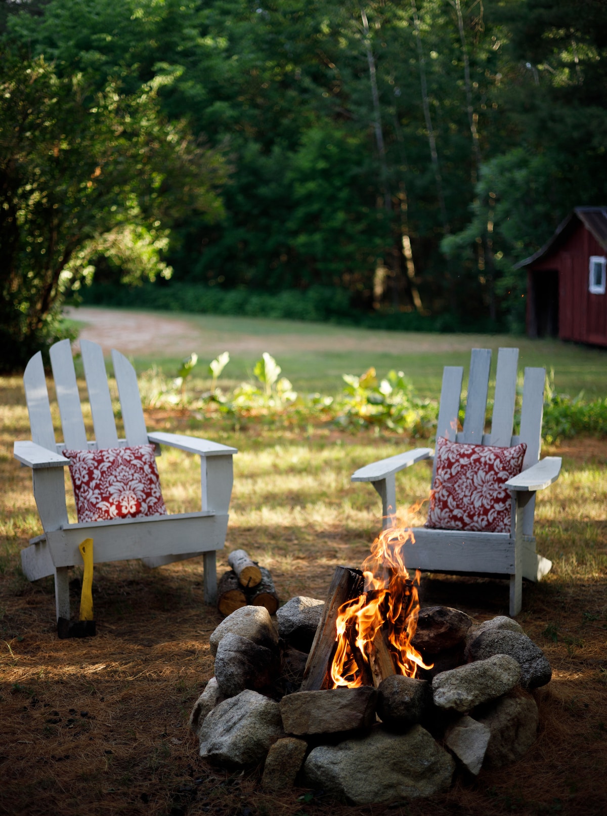 Two white Adirondack chairs with red patterned cushions are positioned around a stone fire pit. Flames reach upward from a cluster of logs inside the pit, set against a backdrop of greenery. A rustic red building is visible in the distance, adding to the outdoor ambiance.