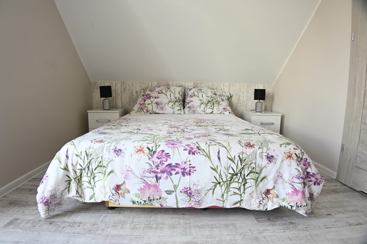 The bedroom features a spacious bed adorned with a floral patterned duvet cover. Bedside tables with lamps are positioned on either side. Light-colored walls and wooden flooring contribute to the airy feel of the room, which is designed for comfort and relaxation.