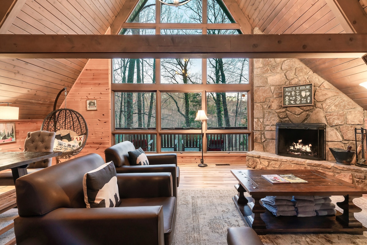 A spacious lounge area features a stone fireplace and large windows that bring in natural light, showcasing forest views. Two leather sofas and a rustic wooden coffee table provide comfortable seating for relaxation. A cozy reading lamp offers a warm glow in the corner.