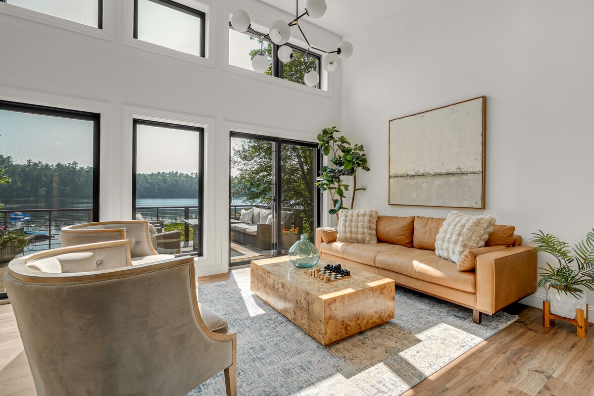 The living area includes a tan sofa with decorative pillows, accompanied by two light-colored chairs. A wooden coffee table is centered on a patterned rug. Large windows provide abundant natural light and views of Hogan Pond, enhancing the inviting atmosphere.