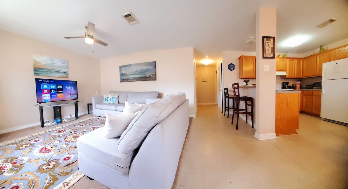 A spacious living area is furnished with a light-colored sectional couch and a large flat-screen TV displaying streaming options. A dining area with high chairs is visible, alongside a well-equipped kitchen with wooden cabinetry and essential appliances. Natural light brightens the neutral-toned room.