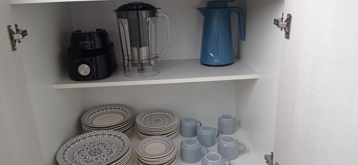 A clean and organized cabinet displays a blender, an electric kettle, and a sandwich maker on the top shelf. The bottom shelf contains a set of patterned dishes and an assortment of light blue cups, providing essential kitchenware for guests.