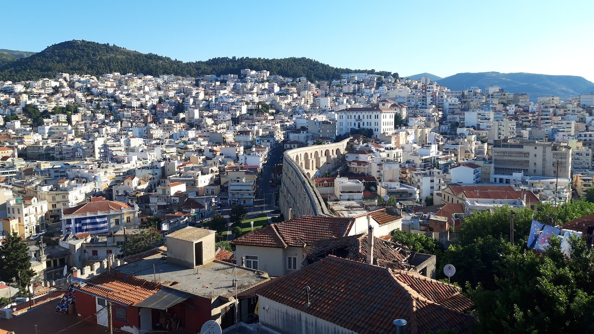 A panoramic view showcases the sprawling city of Kavala, with a blend of traditional and modern architecture. The ancient aqueduct stands prominently amidst the terracotta rooftops. The mountains in the background frame the city, while sunlight bathes the scene, enhancing the vibrant colors of the homes.
