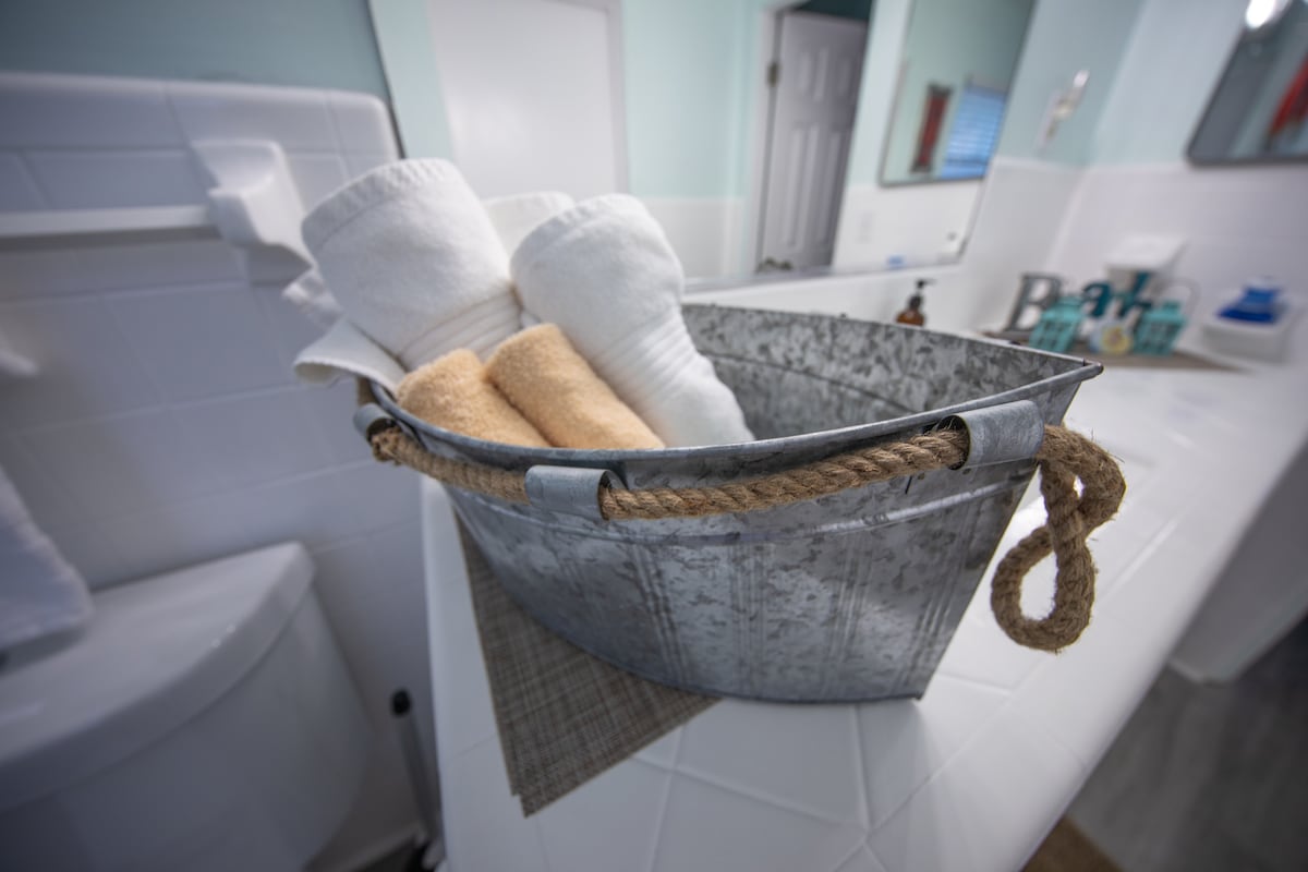 A galvanized metal bucket is filled with rolled white towels and a tan towel, placed on a light-colored countertop. The background includes a mirror and additional toiletries, offering a functional and tidy bathroom setting.