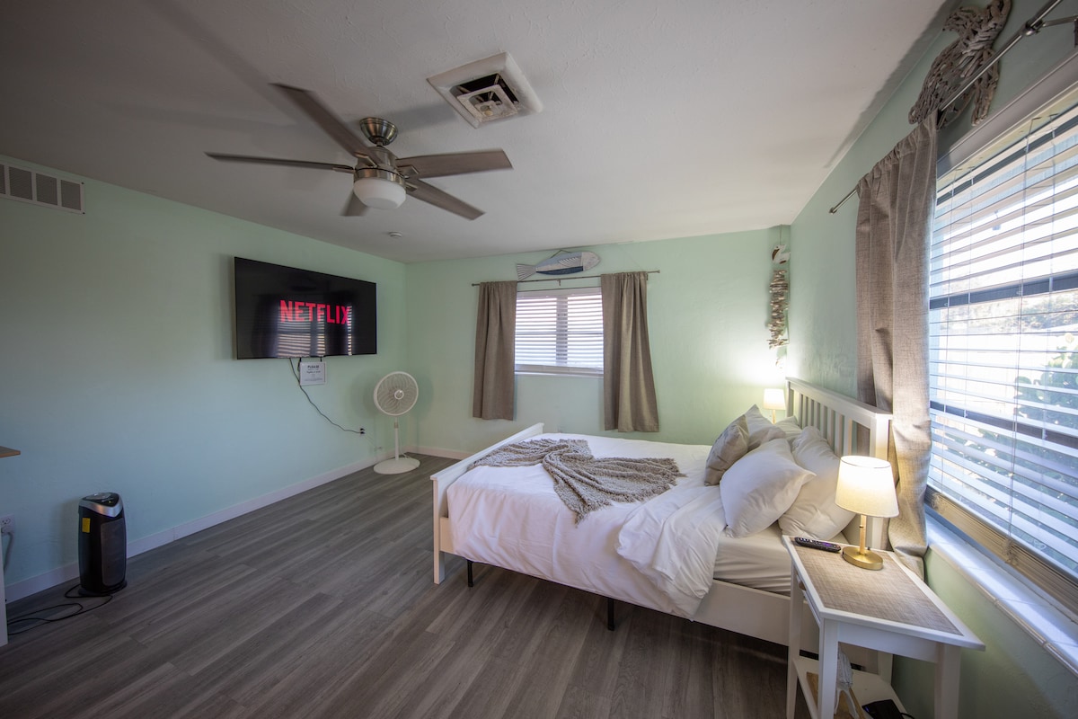 A spacious sleeping area features a queen-sized bed with a light blanket and decorative pillows. A Smart TV displaying Netflix is mounted on the wall, illuminated by natural light from a window. A fan and a small nightstand with a lamp create a relaxing ambiance.