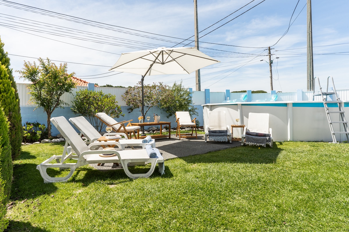 The outdoor area features a well-maintained lawn with several lounge chairs positioned for sunbathing. A large umbrella provides shade over a wooden table. In the background, a swimming pool is seen, with a ladder for access.
