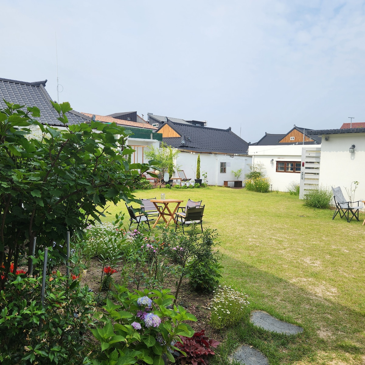 A green lawn is surrounded by blooming flowers and shrubs. Several outdoor seating arrangements, including tables and chairs, are situated throughout the space. Traditional-style buildings are visible in the background under a clear sky.
