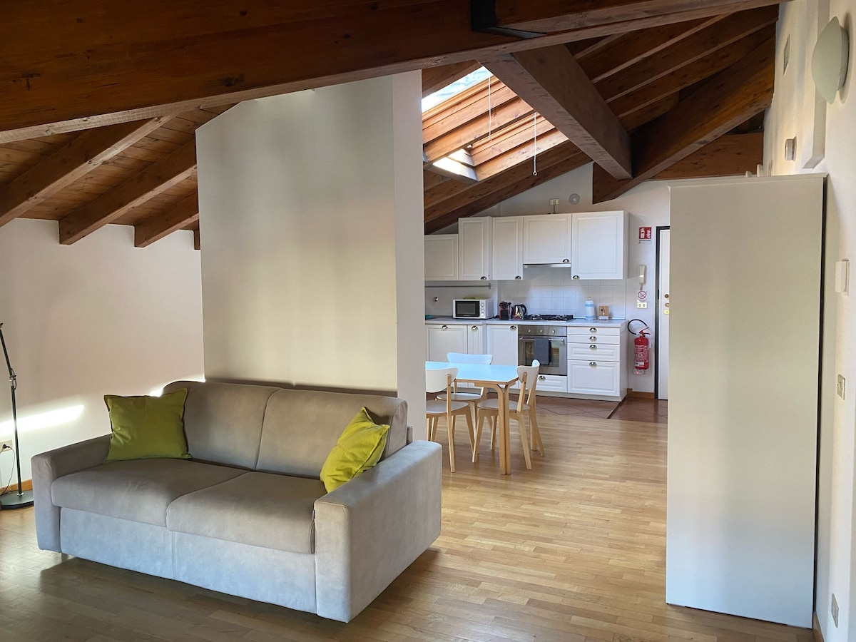 A bright living area features a spacious sofa adorned with green cushions, complemented by wooden beams on the ceiling. An open kitchen with modern appliances is visible in the background, alongside a dining table surrounded by four chairs, creating a cozy and functional space.