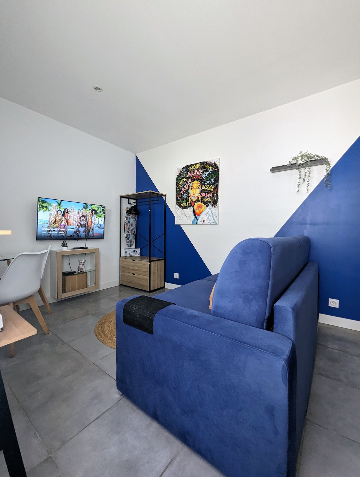 A modern living space features a blue sofa positioned along a wall with a contrasting geometric design. A television is mounted nearby, displaying a vibrant image. Bright natural light enhances the airy feel, while a small shelving unit and a decorative touch add character to the room.