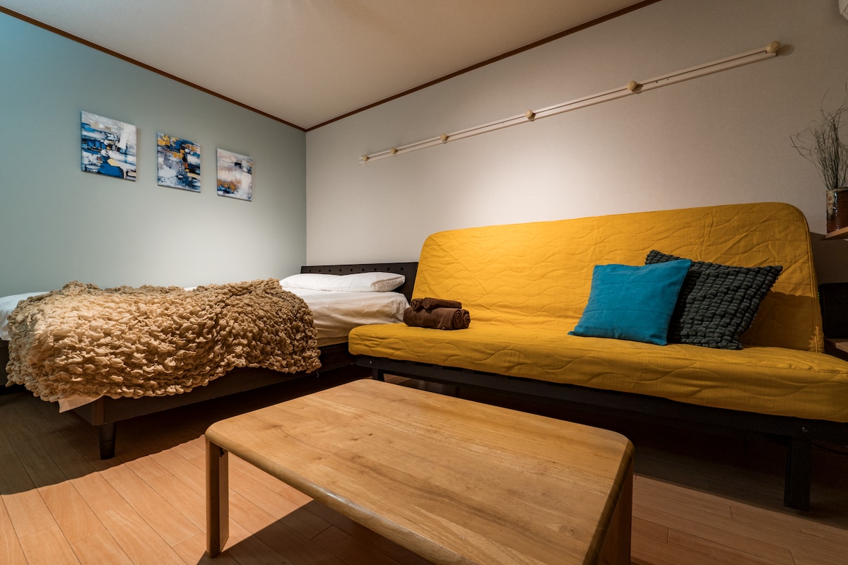 A cozy room is presented with a double bed and a yellow sofa, complemented by decorative cushions. A wooden coffee table is positioned in the center, offering a practical space. Soft lighting highlights the inviting atmosphere, while artwork adorns the walls, enhancing the overall design.