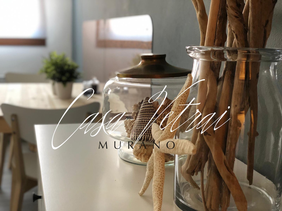 A clear glass jar containing natural elements like starfish and branches is positioned on a light-colored surface. In the background, a simple dining table and chairs are visible, complementing the space's overall ambiance.