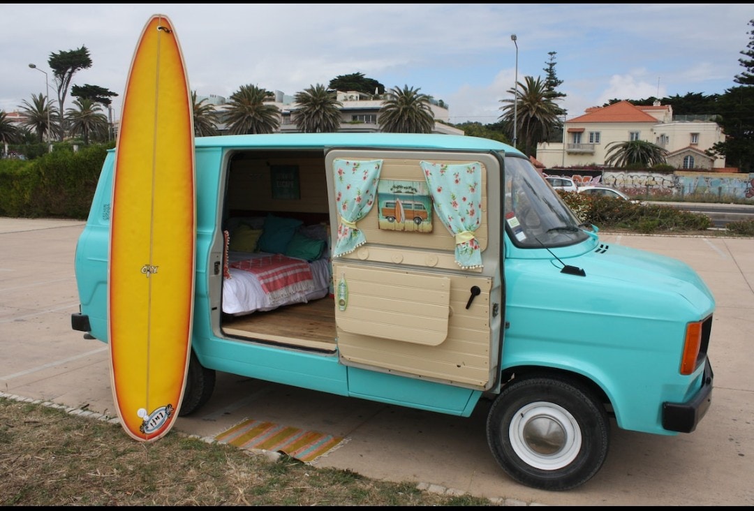 A vintage van in a soft turquoise hue is parked on a grassy area, with a bright yellow surfboard leaning against it. The entrance reveals a cozy interior featuring light bedding and cheerful curtains that enhance the van's inviting character.