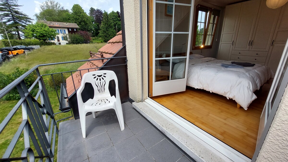A balcony is visible, featuring a white plastic chair. The balcony door is open, revealing a spacious bedroom with two beds and a large wardrobe. A grassy area is seen in the background, along with distant houses and trees, creating a serene setting.