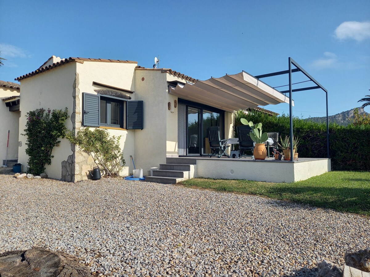 A bungalow is shown with a gravel pathway leading to the front entrance. A covered patio area is visible, surrounded by potted plants. The exterior features light-colored walls and dark blue shutters, inviting natural light into the interior.