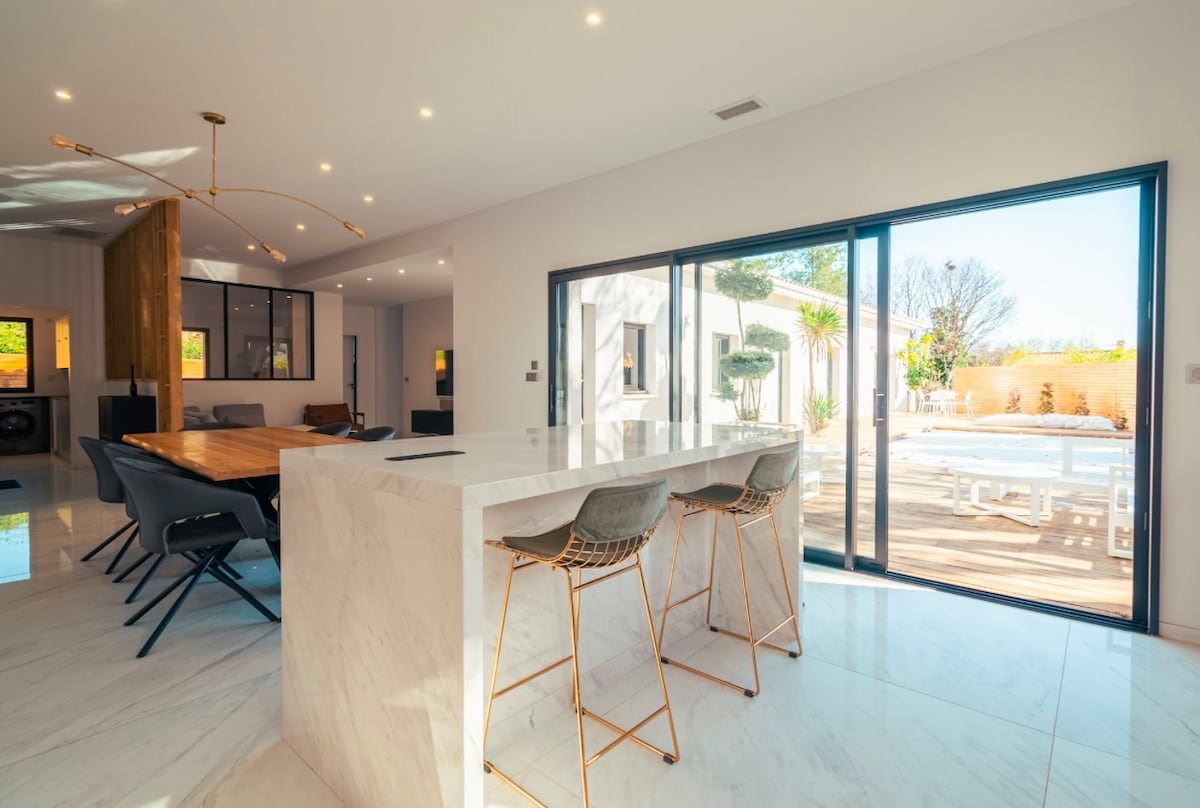 An open-plan kitchen and dining area is featured, characterized by sleek marble countertops and modern bar stools. Large glass doors provide a view of the outdoor pool and deck, allowing natural light to fill the space and create a bright atmosphere.