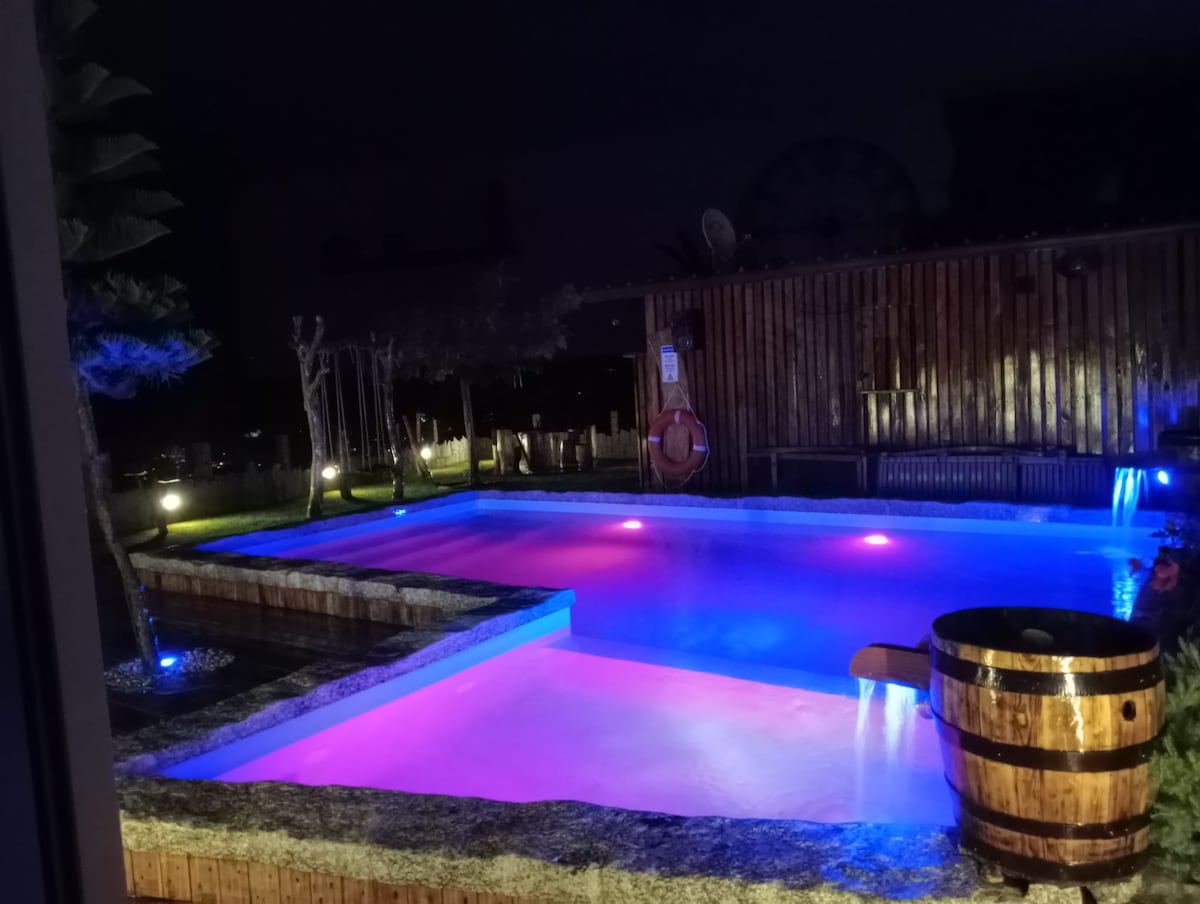 A serene outdoor pool area is illuminated with soft, multicolored lights, creating a calming ambiance at night. The surrounding wooden fencing adds a natural touch, while a wooden barrel is positioned nearby. Lush greenery can be seen in the background, enhancing the private space.