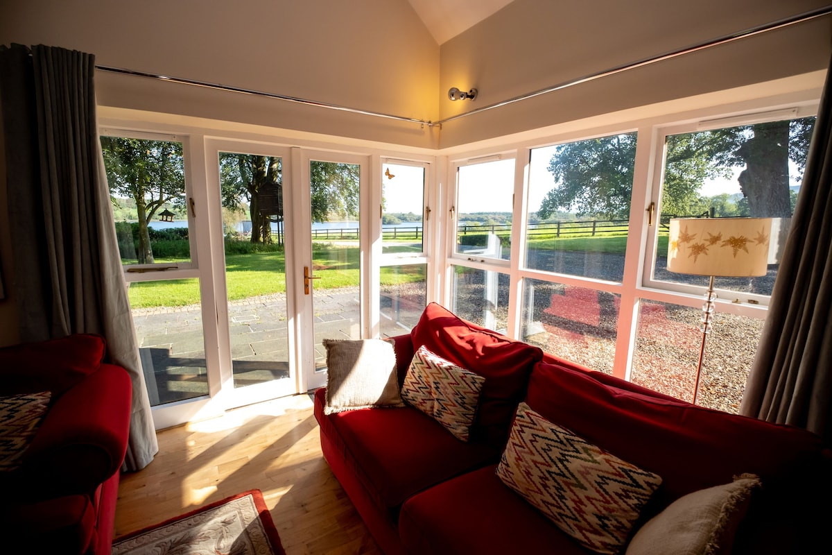A cozy seating area features a red sofa adorned with decorative pillows. Large glass doors allow natural light to fill the space and provide views of the landscaped grounds, including trees and open fields beyond.
