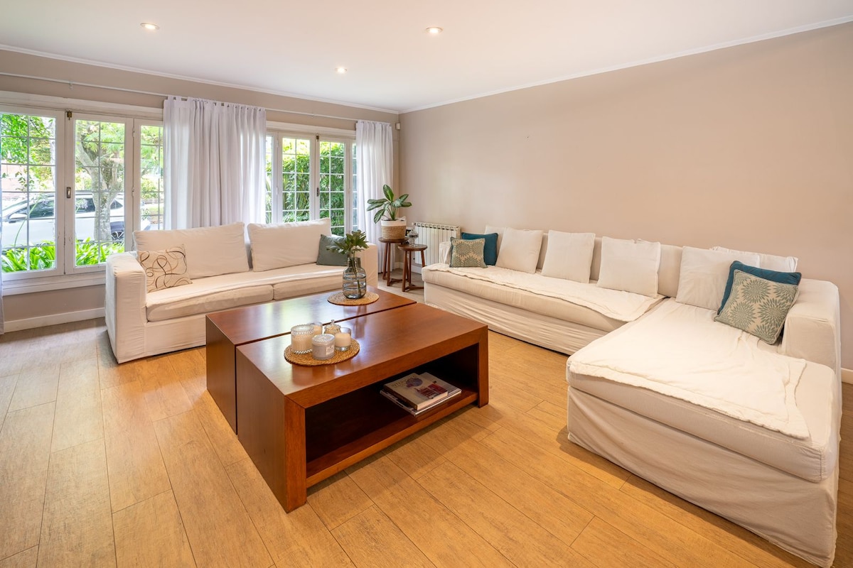 A spacious living area is visible, featuring comfortable sofas and a wooden coffee table. Large windows allow natural light to fill the space, framed by white curtains. A small plant adds a touch of greenery, enhancing the inviting atmosphere of the room.