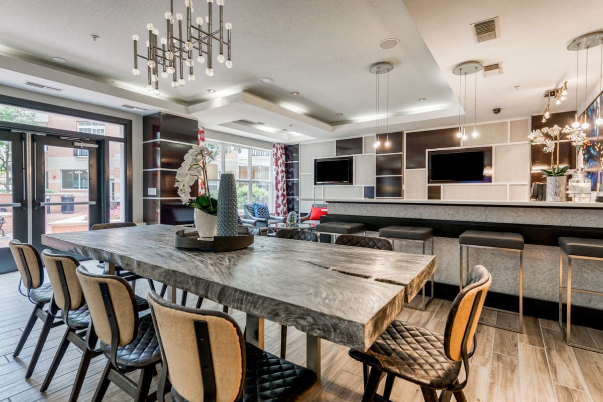 A communal lounge area is depicted, featuring a large wooden table surrounded by several chairs. Vertical floral arrangements are placed on the table, and stylish lighting fixtures hang above. Two large televisions are mounted on the wall, enhancing the space's modern design.