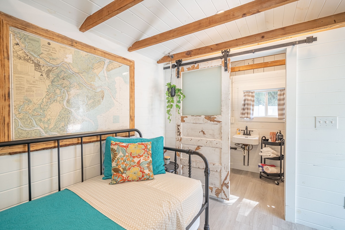 A cozy bedroom showcases a single bed adorned with a colorful pillow, complemented by a framed map on the wall. Sliding barn doors provide access to an adjoining bathroom area, which features natural light and a neutral color palette.
