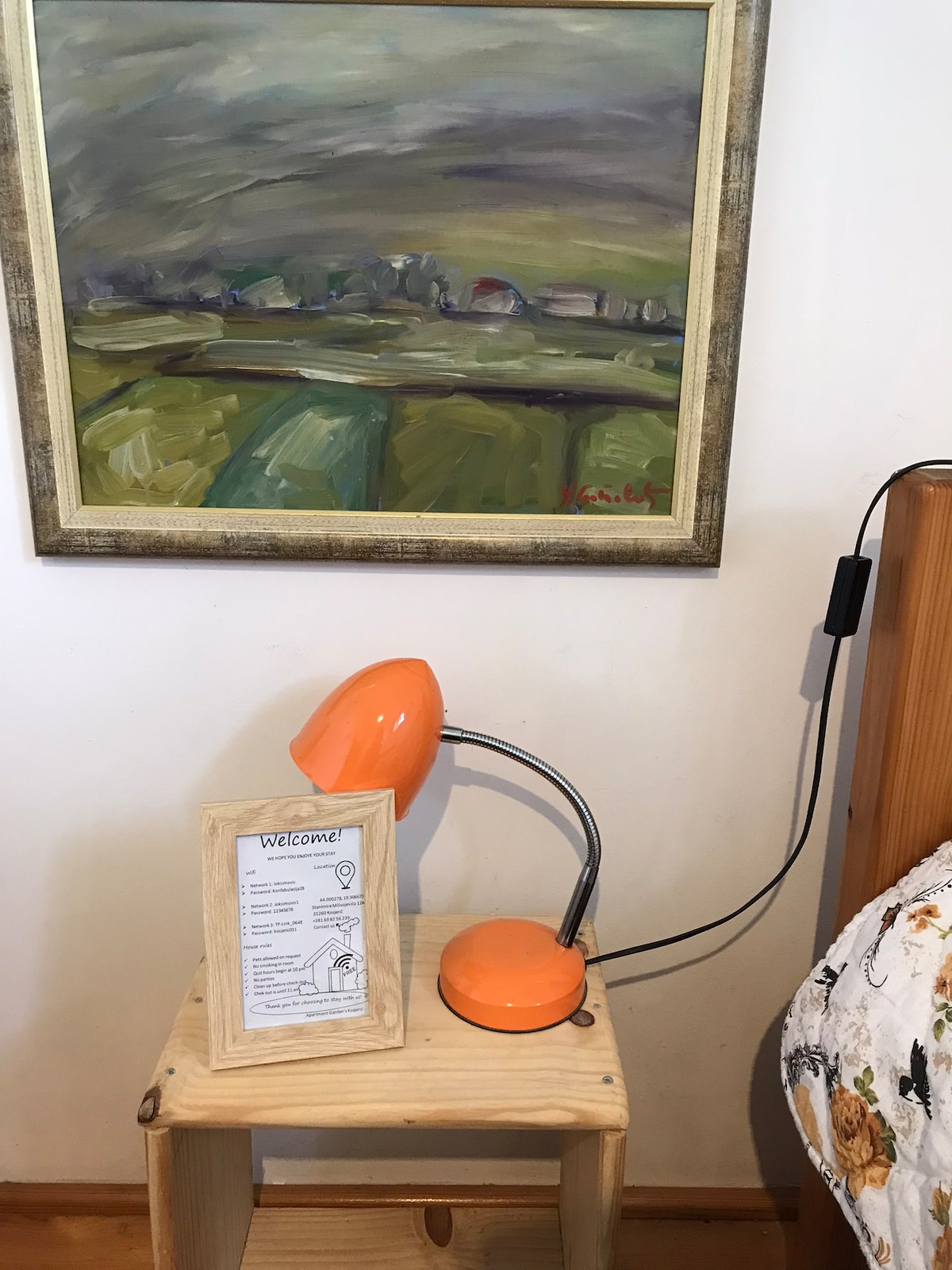 A small wooden nightstand hosts an orange desk lamp, providing a splash of color. Next to the lamp, a welcome note is placed on the surface. Above, a landscape painting in muted colors adds an artistic touch to the room.