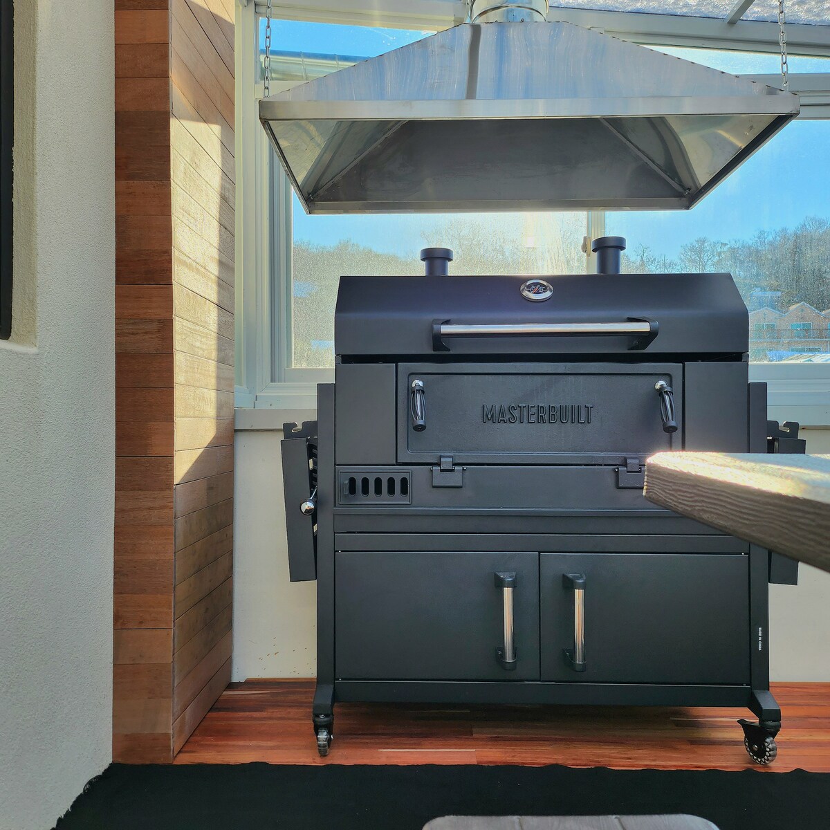 A large, black outdoor grill is positioned under a stainless steel hood within a well-lit space. The grill features two doors and burners, showcasing a functional design. The surrounding area is complemented by wooden paneling and natural light streaming in through a window.