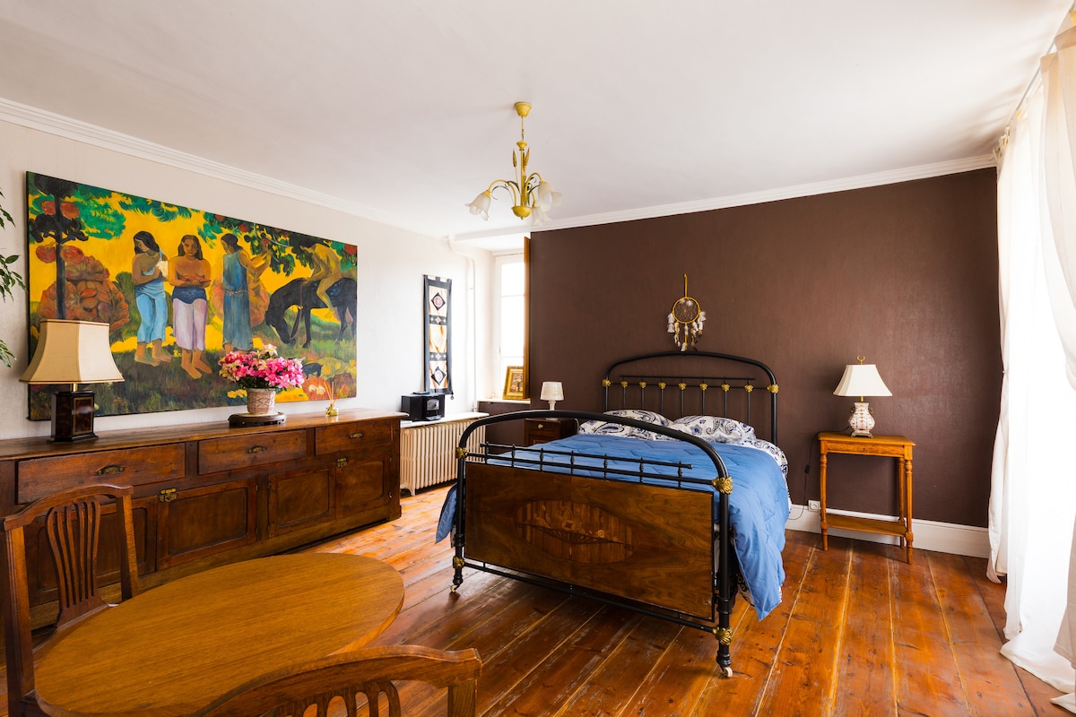 A spacious bedroom is highlighted by a colorfully painted wall art above a wooden sideboard. The room features an antique-style iron bed with a blue coverlet, wooden flooring, and a small dining table. Two lamps illuminate the warm ambiance, complemented by natural light from a window.