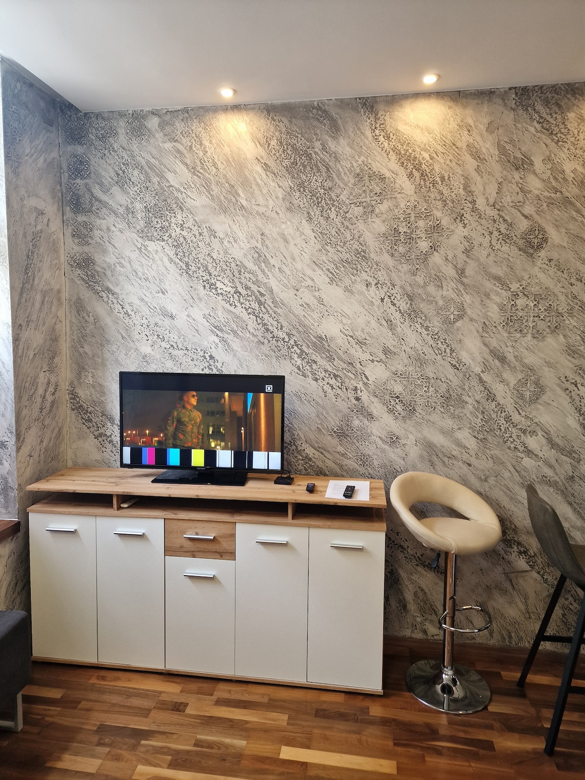 An entertainment area is featured with a flat-screen television positioned on a wooden cabinet. The wall is adorned with a textured design, and subtle lighting emphasizes the space. Nearby seating includes a bar stool and a contemporary chair, enhancing usability.