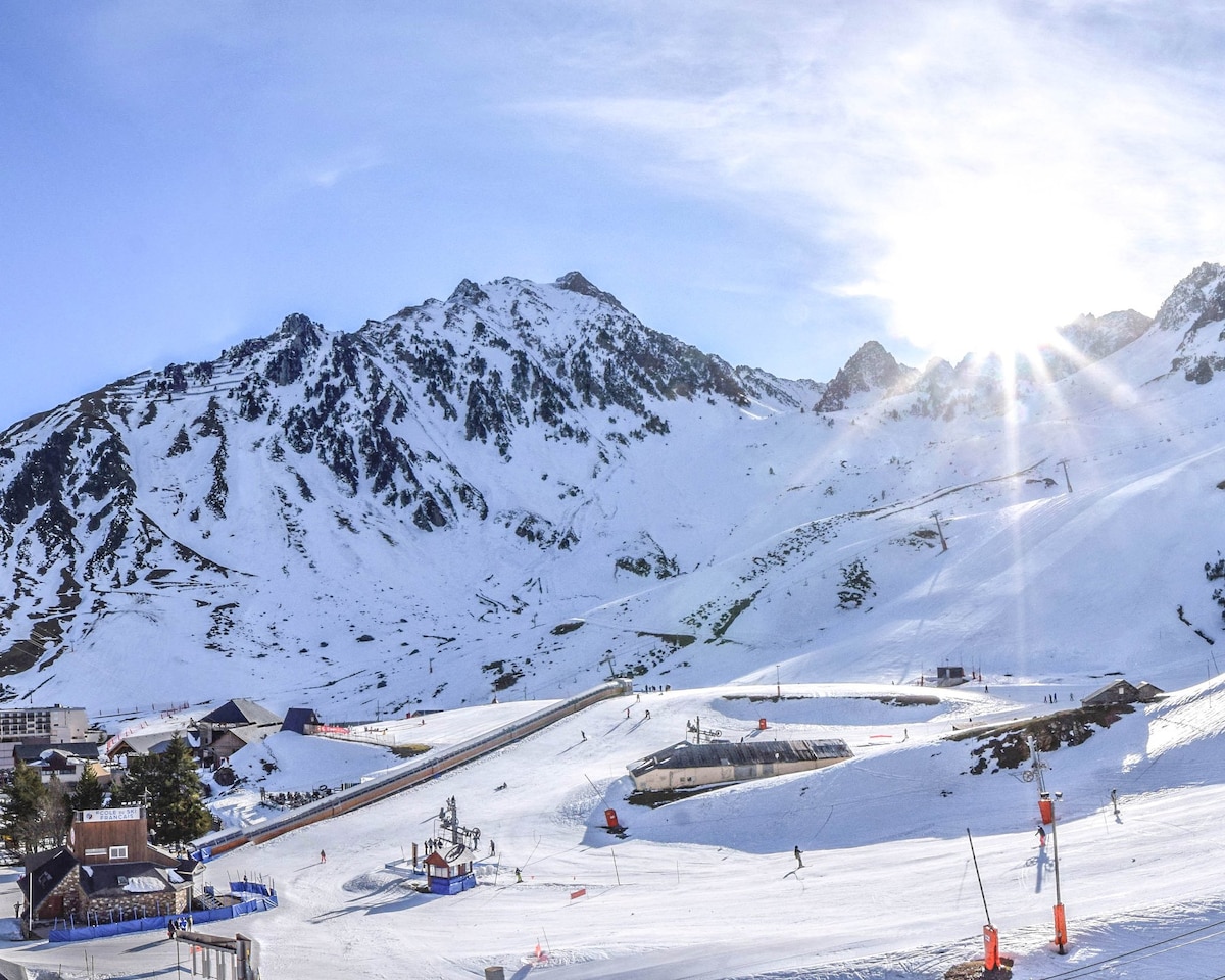 A panoramic view of snow-covered mountains under a clear blue sky, illuminated by sunlight. The slopes are visible, complete with skiers enjoying the terrain. Various buildings and ski lifts dot the landscape, surrounded by a radiant alpine setting.
