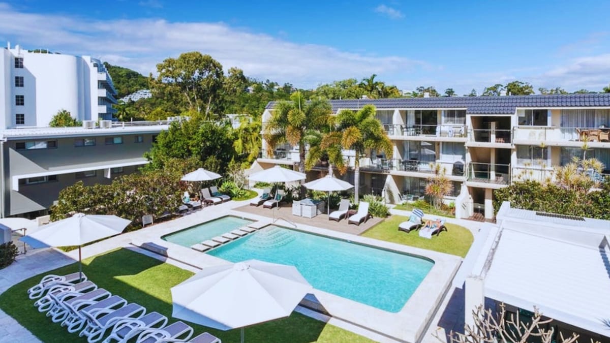 A modern hotel complex features a large heated pool surrounded by spacious sun loungers and shaded umbrellas. Lush greenery enhances the inviting outdoor space. The balconies of the adjacent buildings are visible, providing a sense of relaxed resort living in a vibrant setting.