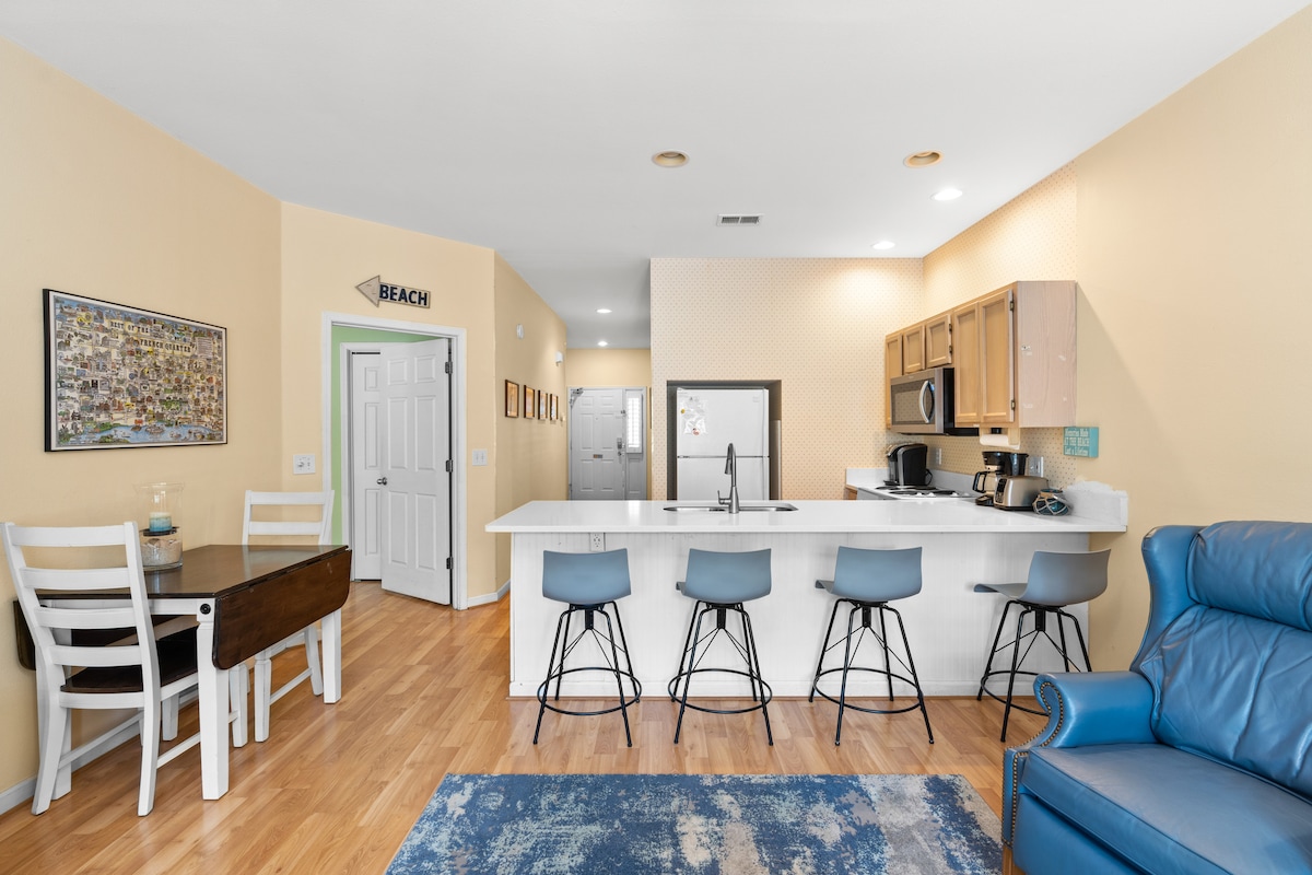 An open living space features a modern kitchen with wooden cabinetry and a central island equipped with bar stools. A small dining table is visible alongside a comfortable blue recliner. Light-colored walls contribute to a bright and airy atmosphere.
