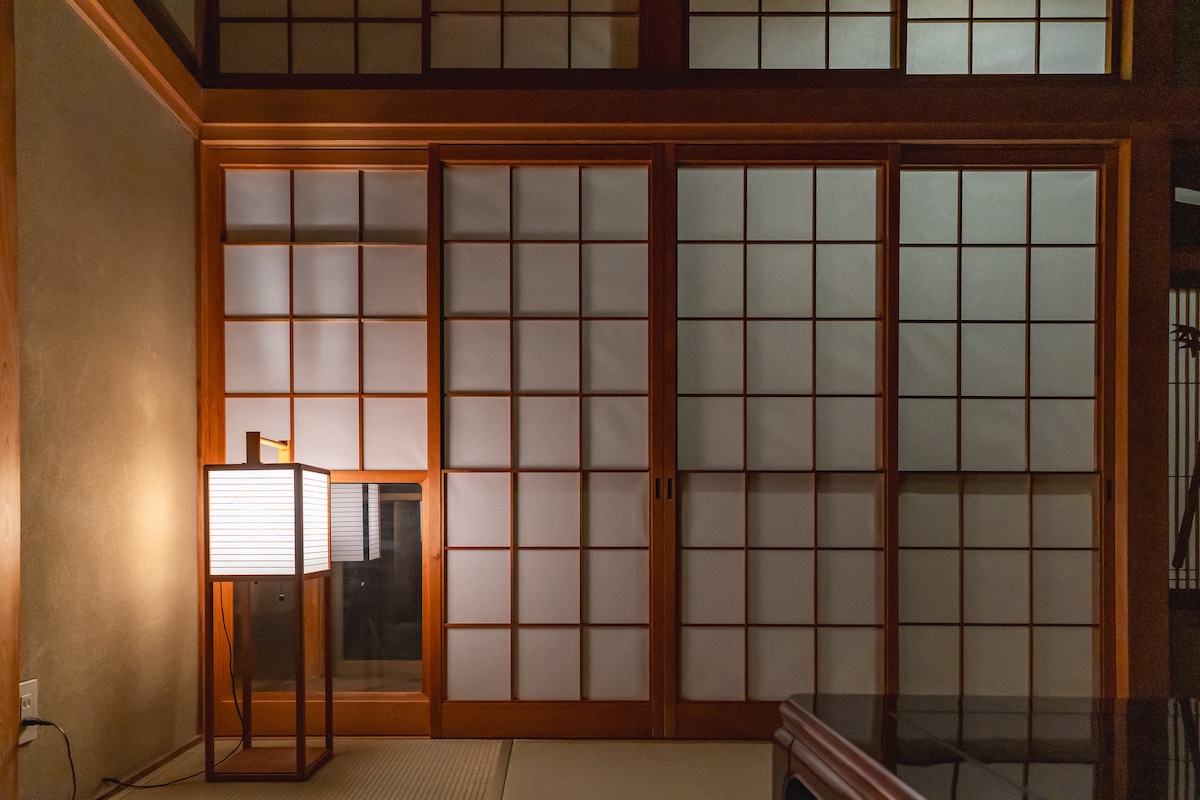 A serene interior is depicted, featuring shoji screens that divide the space and allow soft light to filter through. A warm lamp stands beside the screens, casting a gentle glow. The natural wood framework adds to the tranquility of the environment.