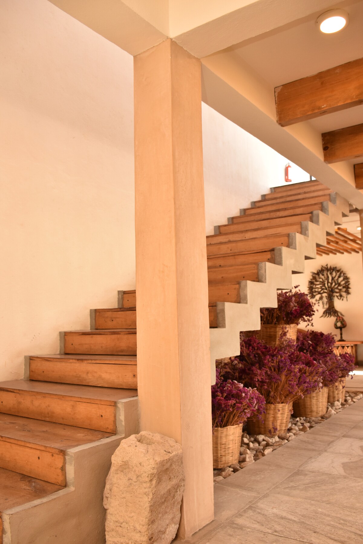A modern staircase is framed by a minimalist design, featuring wooden steps and a concrete structure. Lush purple plants are displayed in woven baskets along the side, enhancing the natural aesthetic. Soft lighting illuminates the space, creating a welcoming atmosphere.