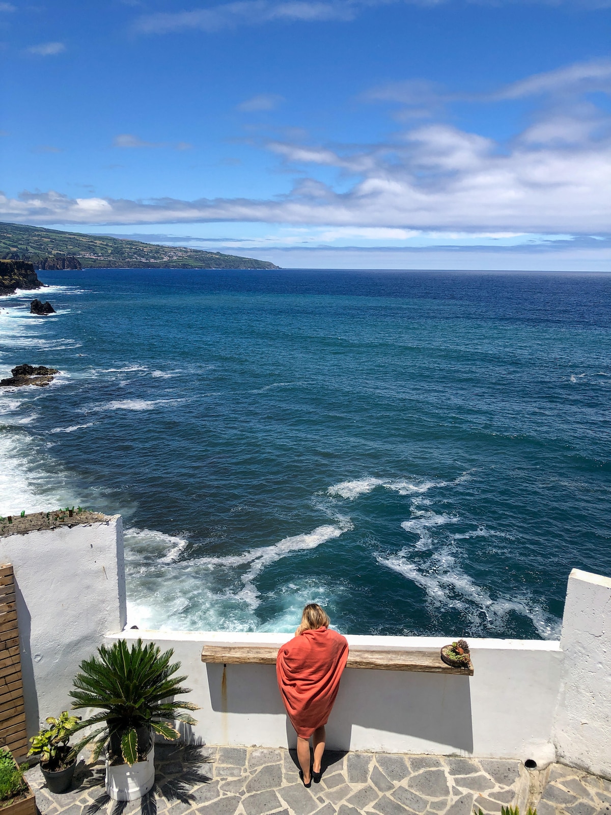 A coastal view is presented, with waves gently breaking against rocky shores. A person stands near a stone wall adorned with a potted plant, gazing out towards the expansive ocean under a bright blue sky interspersed with clouds.