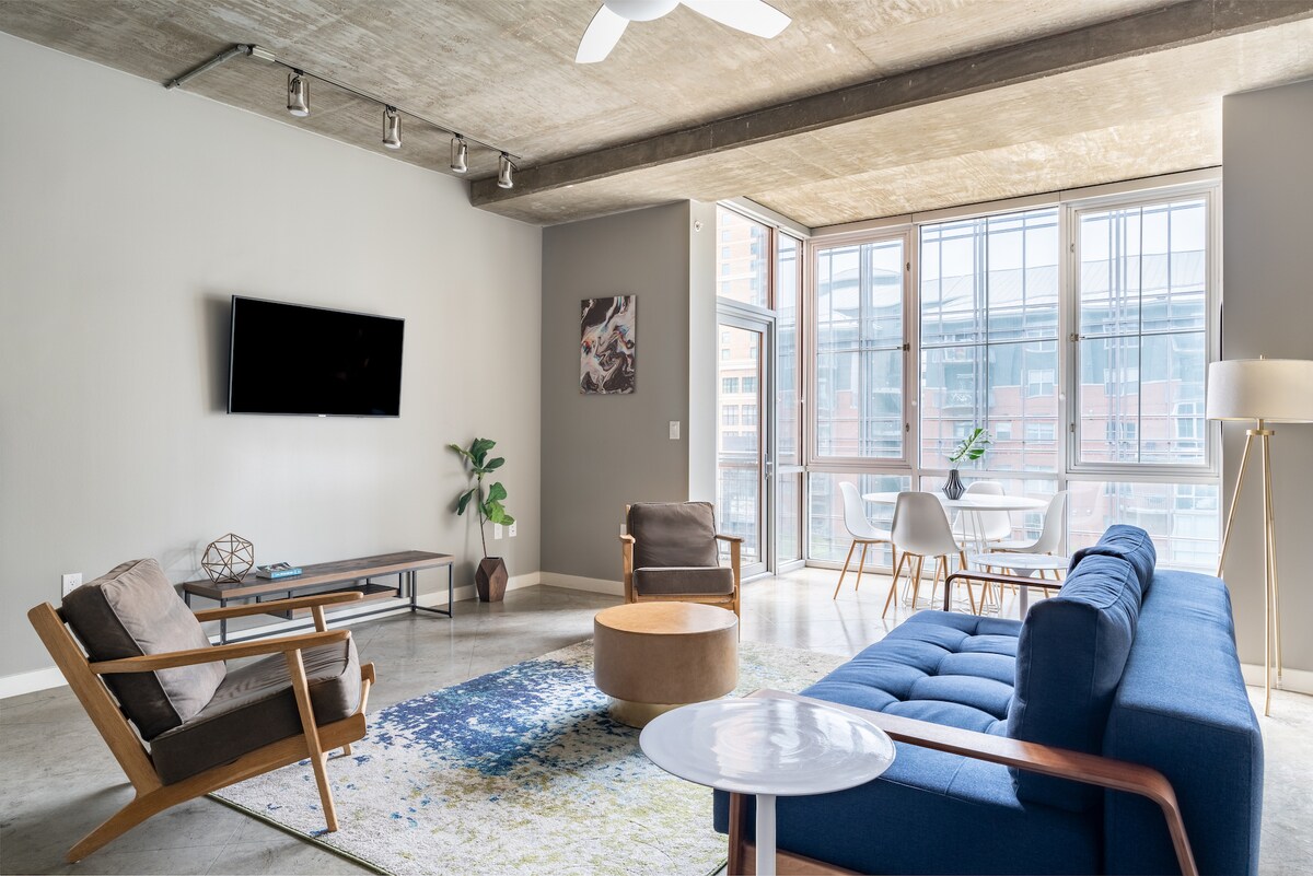 The living space features large windows allowing ample natural light, showcasing a modern design. A blue sofa is complemented by wooden chairs and a round coffee table, while a smart TV is mounted on the wall. A small dining area is visible near the windows.