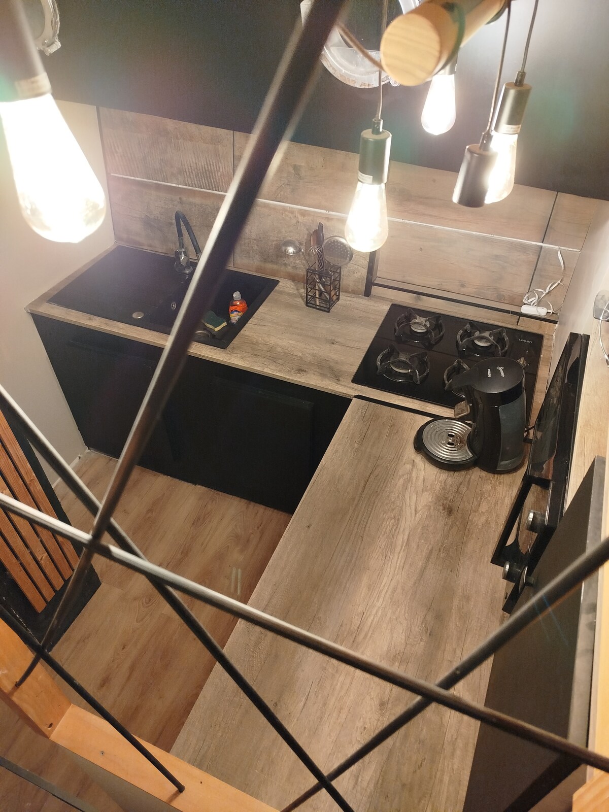 An overhead view of a modern kitchen features a black sink and a gas stovetop set against a wooden backsplash. Ambient lighting is provided by hanging bulbs. A coffee maker and decorative items complement the functional space, which is organized and inviting.