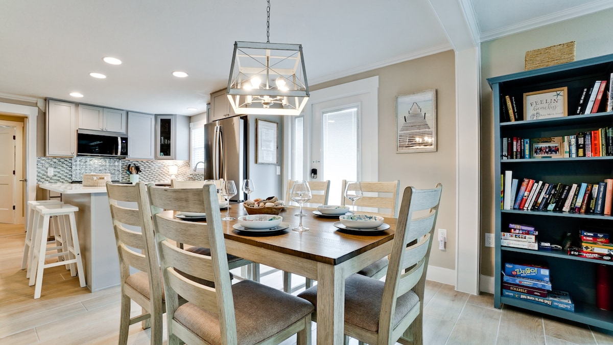 A modern dining area is accented by a wooden table surrounded by six chairs. A light pendant hangs above the table, illuminating the space. In the background, a well-equipped kitchen features sleek cabinetry and stainless-steel appliances, while a bookcase showcases a collection of books.