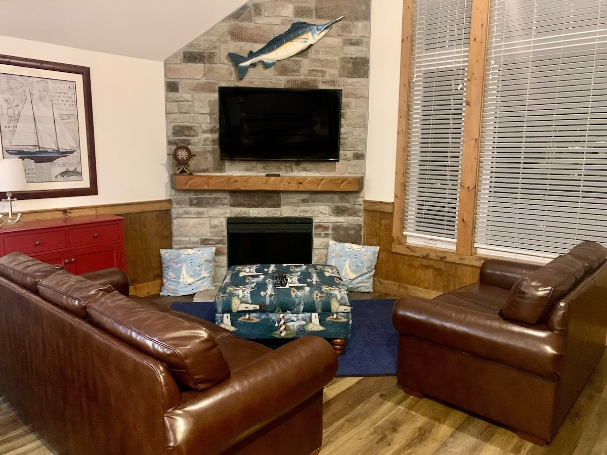 A cozy living area is highlighted by a stone fireplace and a mounted fish decor above a flat-screen TV. Two brown leather armchairs and an upholstered ottoman provide comfortable seating, complemented by light-blue accent pillows. Large windows allow natural light to fill the space.